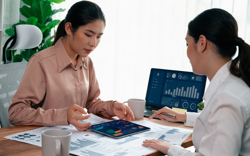 two women at desk analyzing data on papers and iPad