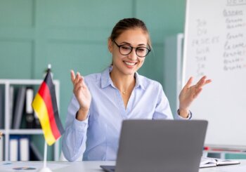 woman in glasses talking on virtual call with german flag on desk and whiteboard behind her